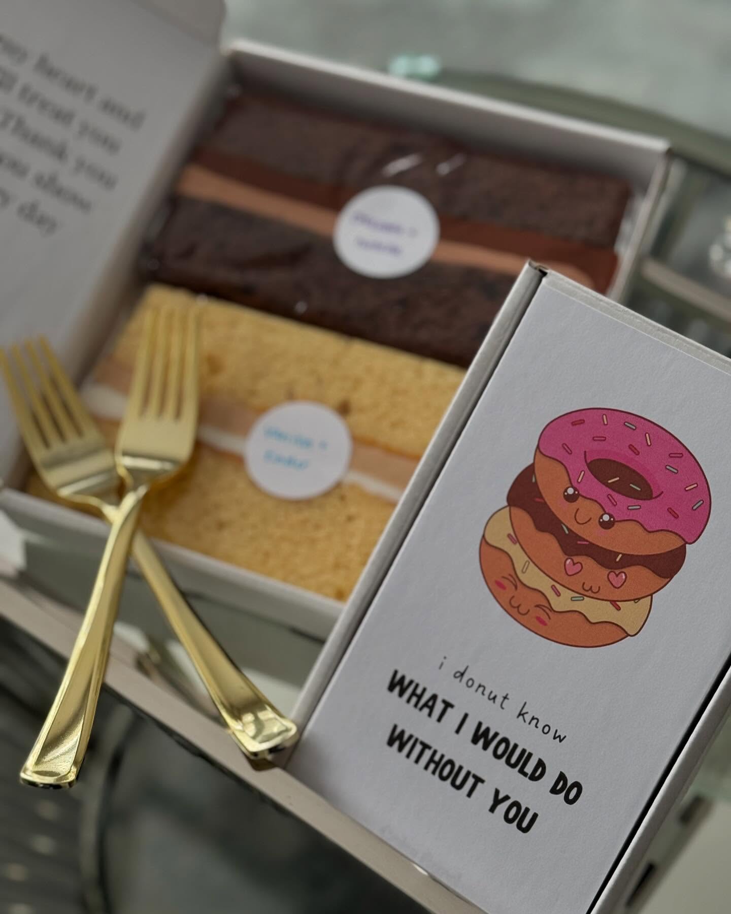 Two slices of layer cake boxed for delivery with gold forks and a donut card